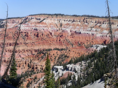 [View looking across the ampitheater in which both the top and bottom are visible across the huge expanse. There are only a scattering of trees at several of the lower levels. The upper levels are shades of white, tan, red, and brown. There is a row of trees long the top ridge.]